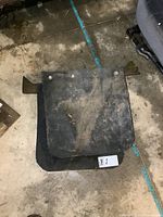 Pair of black rubber mud flaps, square shape with rounded corners, showing dirt and wear, each with an attached steel mounting bracket with holes for installation, placed on a concrete floor near a vehicle seat.