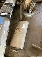Photo shows the entire muffler with pipe and flange laying on a concrete floor. Rust is visible on the muffler's surface.