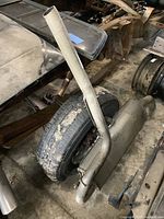 Photo showing the full muffler with attached curved pipe next to a tire and various vehicle parts, with visible wear and surface dirt.