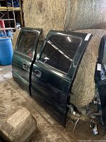 Two dark green full size Chevy truck rear doors leaning against hay bales in a barn. Doors have tinted windows, visible slight surface scratches and light rust near the bottom edges.