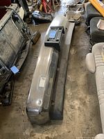 Chrome Chevrolet truck bumper with integrated spoiler and valance shown lying on concrete floor with adjacent vehicle seats and parts visible.