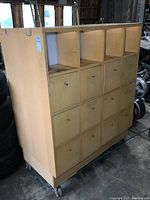 Front view of the large double-sided parts cabinet showing 12 drawers and 4 open cubbies on one side, light wood finish and mounted on casters.