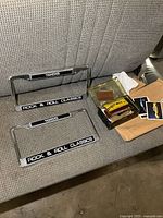 Pair of chrome license plate frames with '1955' and 'ROCK & ROLL CLASSICS' plaques and a small box with car show plaques on a bench seat.
