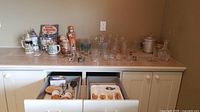 Wide view of glassware on a countertop including clear drinking glasses, mugs, two metal bar tools, and boxed kitchen accessories. Some glasses have logos or green tint.