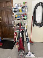 Photo showing red Bissell carpet cleaner and white/blue Bissell steam mop standing on floor with cleaning supplies on wall rack behind them.