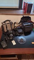 Overview of two camera bags, Nikon SLR body with lens, flash pouch, camcorder in open bag, binoculars, film reel and charger on desk