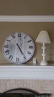 White ceramic table lamp with floral base and cream pleated fabric shade next to large round wall clock with Roman numerals on distressed white face, sitting on fireplace mantel
