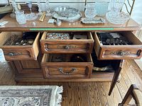 Photo of a wooden sideboard showing four partly open drawers and one open cupboard door revealing silver plated cutlery and serving items inside. Several silver plated trays and dishes displayed on top of the sideboard.