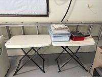 Two white plastic folding tables with black metal legs side by side with items on top in a garage setting.