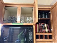 Wide view showing upper kitchen cabinets with blue and clear textured glass wine glasses, vintage storage jars, and assorted cookbooks on wooden shelves above a microwave oven.