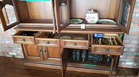Wooden hutch cabinet with drawers open showing silverware in trays and several clear glass wine glasses hanging from racks below the drawers