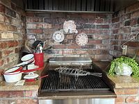 Wide view of kitchen countertop showing red and white enamel pots, utensils in red holder, wire rack on a grill and three decorative plates on brick wall