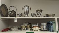 Shelf containing silver on copper dishware including kettle, samovar urn, sugar bowls, small serving dishes, and various bakeware items plus a tin container and ceramic dish in bottom right corner.