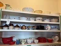 Wide view of shelves containing multiple bowls, ceramic jugs, mugs, kitchen scale, and jars.