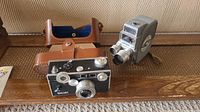 Two vintage cameras shown including the black Argus film camera with dials and leather case, and the silver-gray Keystone 8mm Electric Eye K-4C movie camera.
