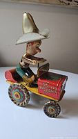 Side view of Rodeo Joe tin toy showing cowboy figure with large silver hat on red jeep marked 'Git Along Lil' Doggie' with colorful lithographic wheels.
