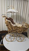 Side view of antique wicker baby stroller showing intricate wicker weaving, metal wheels, and lace-trimmed white parasol.