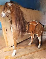 Side and front view of antique horse toy made of wood covered in hide on burlap, with detailed mane and leather tack, placed on wooden slats missing the base.