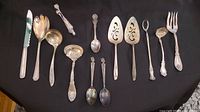 Full view of the eleven antique and vintage silver plated Rogers flatware pieces arranged on black cloth, showing various shapes including serving spoons, forks, ladle, and knife with intricate detailed patterns and some tarnishing.