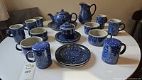 Photo showing full set of blue spackle-speckled ceramic items including teapot, cups, plates, shakers, sugar dish, and pitcher arranged on table.