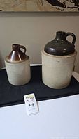 Two antique stoneware jugs, beige bases with glazed brown tops, displayed together on black surface.