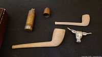 Photo showing two straight stem pipes, one with a carved clover pattern, WW1 lighter with brass casing, and silver-tone bull head bolo tie clasp.