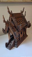 Side view showing the intricate roof details, stairway, and front door of the wood carved Thai spirit house.
