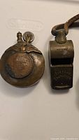 Photo showing both the antique brass trench lighter and Acme Thunderer whistle side by side on a white background.