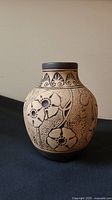 Front angle showing carved floral design on rounded beige pottery vase with dark patterned neck band.