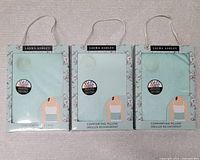 Three Laura Ashley light turquoise boxes each containing one comforting pillow, arranged side by side on a light carpeted floor.