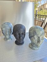 Front view of three styrofoam mannequin heads, two with blue-gray collage finish and one black, placed on a table outdoors with background foliage.