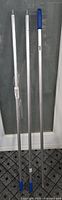 Three brand new industrial aluminum broom handles standing upright, two with threaded ends and one with blue plastic grip.