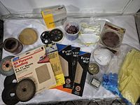 Photo of assorted sanding sheets, Velcro sanding backing pads, brown abrasive discs, and small abrasive discs in container, including original packaging of some items