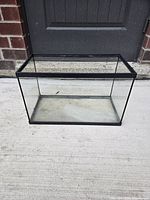 Glass aquarium tank sitting on concrete beside brick wall and door. Clean and transparent with black trimmed edges.