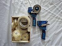 Photo of two blue tape dispensers and a box with multiple rolls of clear plastic tape, both loose and wrapped, on a white surface.