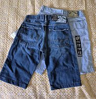 Two pairs of jean shorts, one dark blue denim and one light blue denim, laid out flat showing back sides and brand labels.