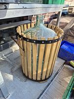 Clear glass demijohn inside cylindrical wooden slat frame with metal handles, photographed outdoors on concrete beneath metal table.