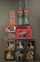 Photo showing entire Coca-Cola collectibles lot including 2 glass bottles, coasters, collector tin with playing cards, matchbox toy car and some pins/keychains