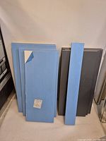 Nine MDF boards shipped stacked vertically: blue protective plastic film mostly intact showing white finish underneath; some boards have grey finish visible on the reverse side.