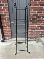 Full view of tall black metal shelf unit with three glass shelves, decorative curled ends on vertical supports.