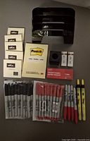 Photo showing assortment of office supplies laid out on a dark surface, including multiple Sharpie markers (black, red, yellow), Offix sticky notes, Post-it notepad, a box of staples, erasers, and black plastic three-tier desk organizer trays.