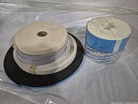 Photo showing multiple large rolls including white fabric strip, white vinyl tape, and double-sided tape with blue protective lining, arranged on a surface
