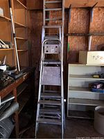 Full view of 5-foot aluminum step ladder placed upright against shelving in a shed, showing three steps and top platform, aluminum construction with wear and paint marks