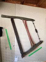 Full view of antique wooden framed buck saw hanging on pegboard showing wooden frame, red tension arms, twisted wire tension, and large toothed metal blade.