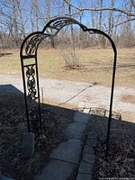 Front view of the black metal garden arbor set along a paved path with trees in background