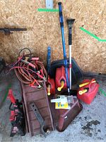 Lot of automotive items including jumper cables, gas cans, car ramps, jack stands, and ice scrapers arranged on a concrete floor against a wooden wall.