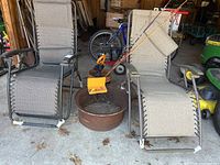Two foldable lounge chairs beside round metal fire pit with grill grate, fishing rods leaning against chairs, small orange fishing net in fire pit.