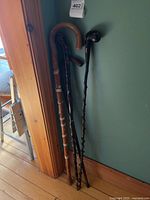 Four walking aids leaning against a wall: two thin dark wooden sticks with twig-style texture, and two light-colored wooden canes, one with wear patches.