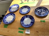Five Flow Blue china plates displayed on wood table with handwritten labels identifying Myotts Crumlin (2), Wood and Son Seville (2), and Lahore (1) patterns.