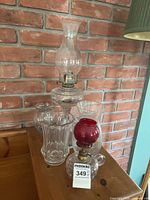Overall view of the lot showing two oil lamps and three clear glass vases against a brick wall background